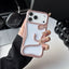 A hand holding a white iPhone case with a metallic rose gold snake design wrapping around the back, set against a desk background with a laptop and headphones.