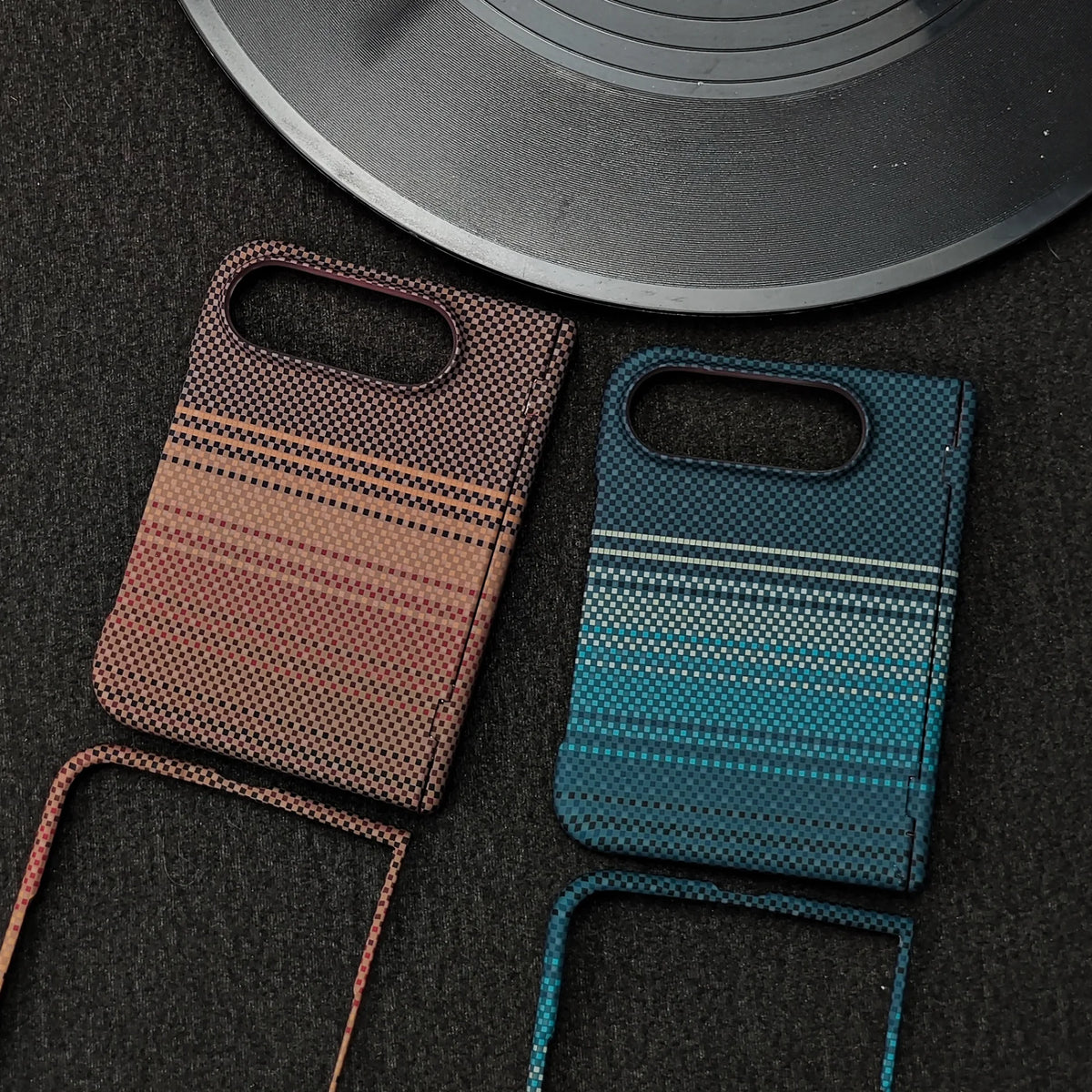 Two patterned phone cases with matching lanyards, displayed on a dark textured surface next to a vinyl record. One case has a rust-orange and brown gradient with black dotted stripes, and the other has a teal and blue gradient with white dotted stripes.