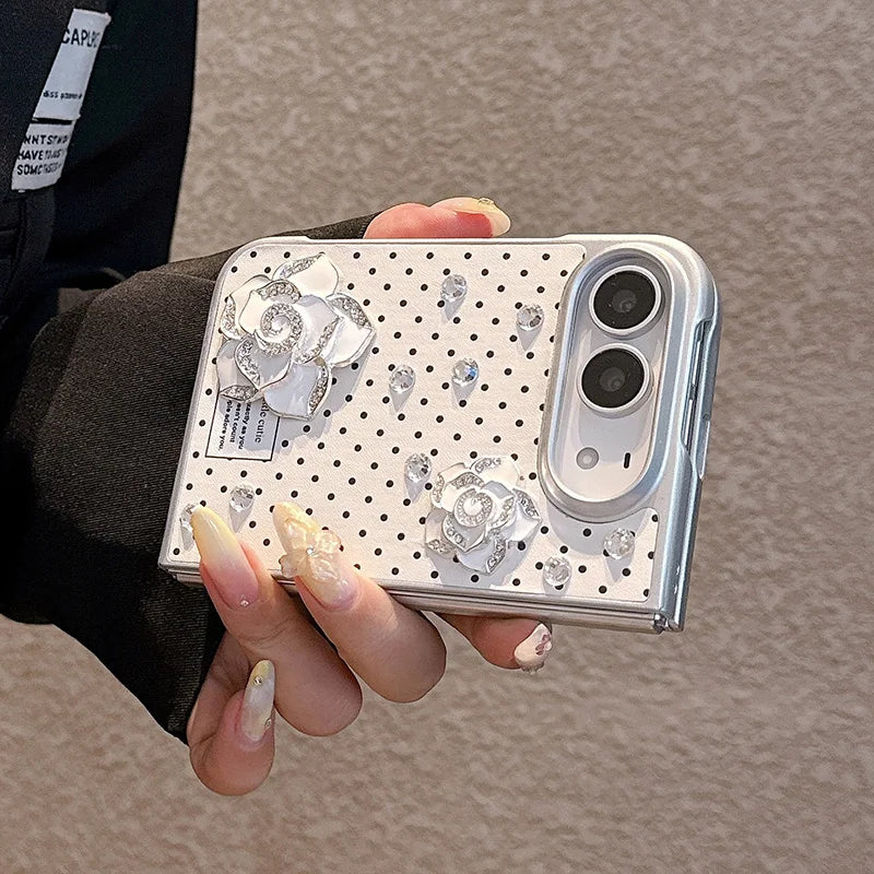 A hand with long manicured nails holds a silver smartphone in a clear case decorated with white rose embellishments, rhinestones, and a polka-dot pattern, set against a neutral textured background.