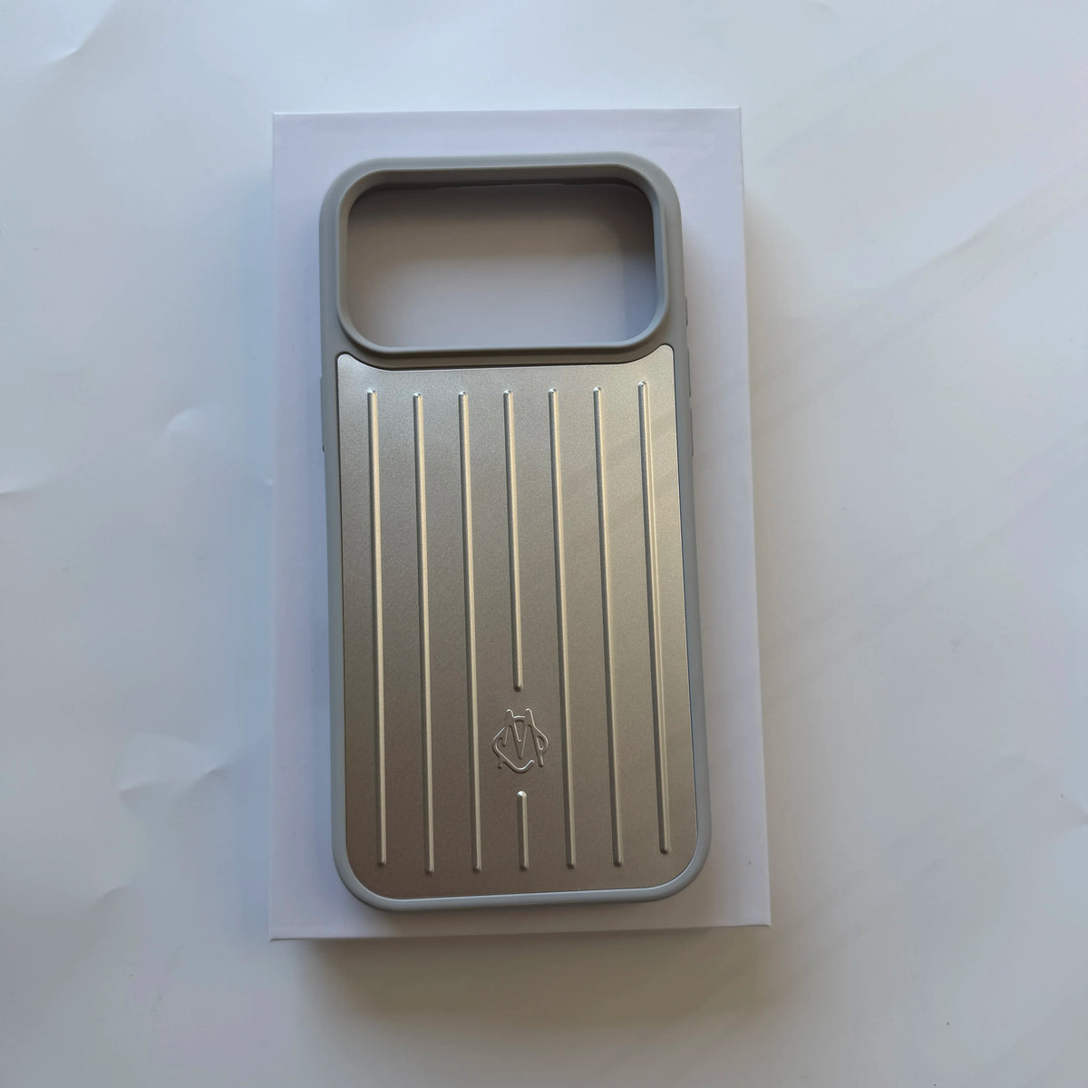 A metallic silver phone case with a vertical ribbed design and a small embossed logo, displayed on a white backing card against a light gray background.