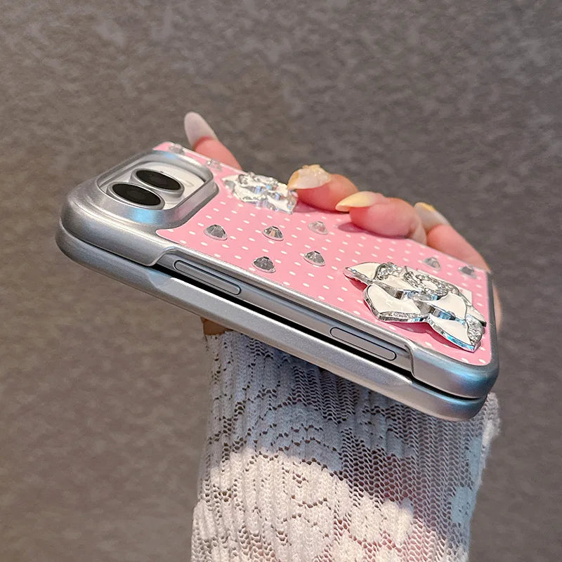 A close-up of a hand holding a pink phone case decorated with rhinestones, small heart patterns, and a large silver crystal bow. The hand is wearing a white lace sleeve, and the background is a textured gray wall.
