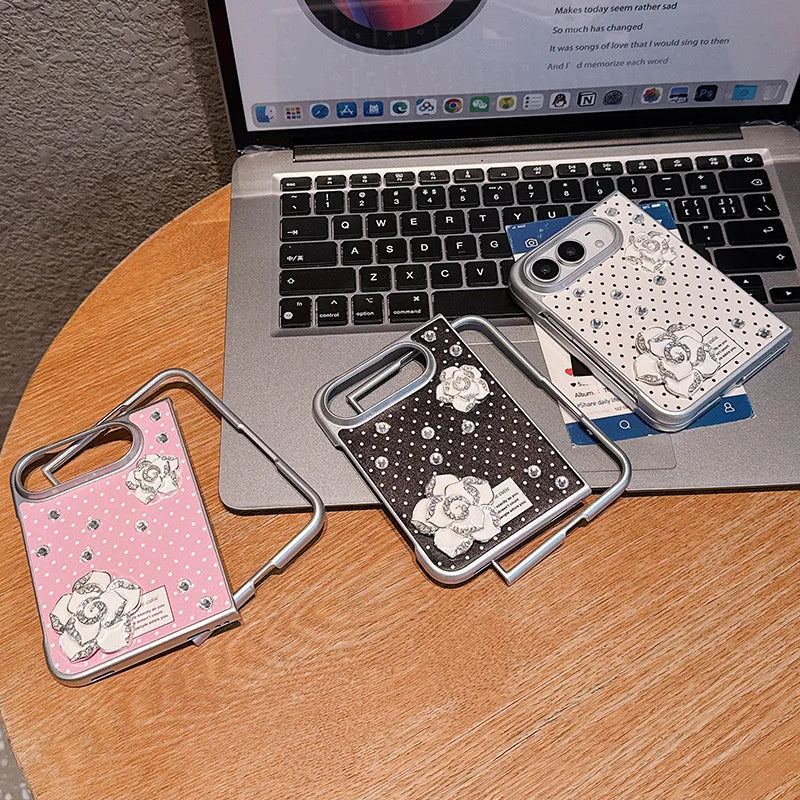 Three decorative phone cases with metal stand arms are arranged on a wooden table in front of an open laptop. The cases feature floral and polka dot designs in pink, black, and white, with one case showing a visible brand label that reads 'Camellia'.