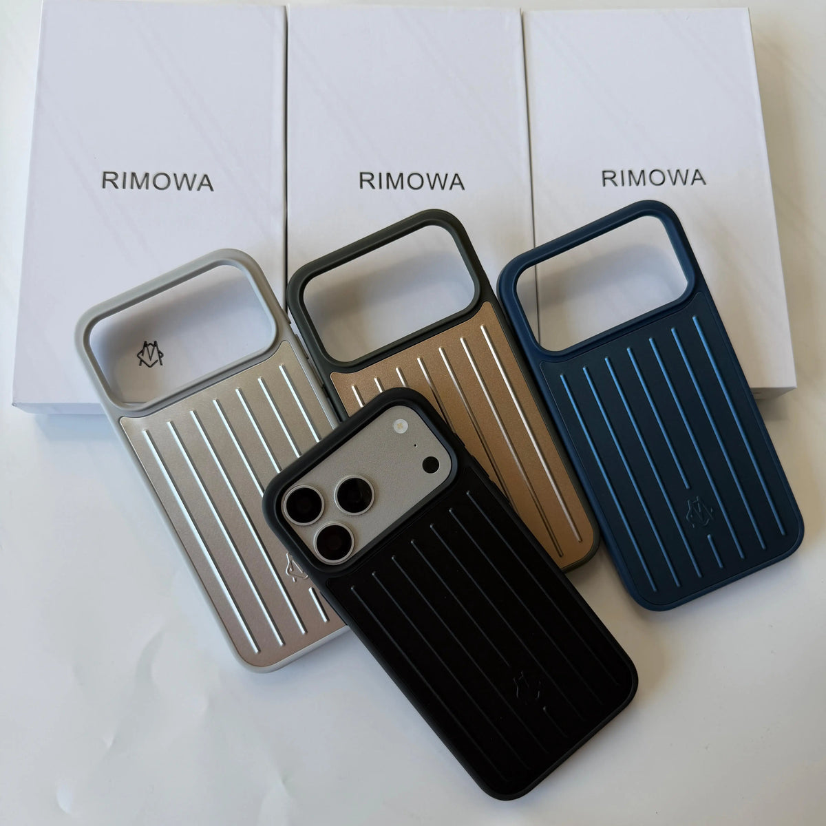 Three slim phone cases with a ribbed rectangular back and camera cutout are arranged in front of three white Rimowa boxes. The cases are shown in silver, bronze, and dark blue, with one black case placed in front.