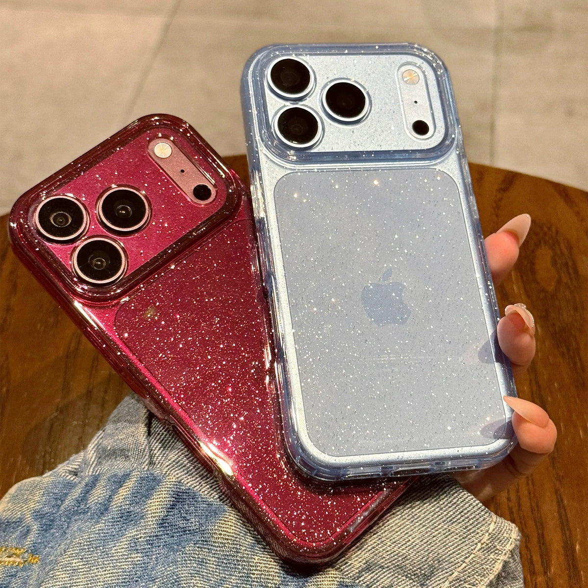 Two glittery phone cases held in a hand over a wooden table. One case is metallic rose gold/magenta and the other is light blue.