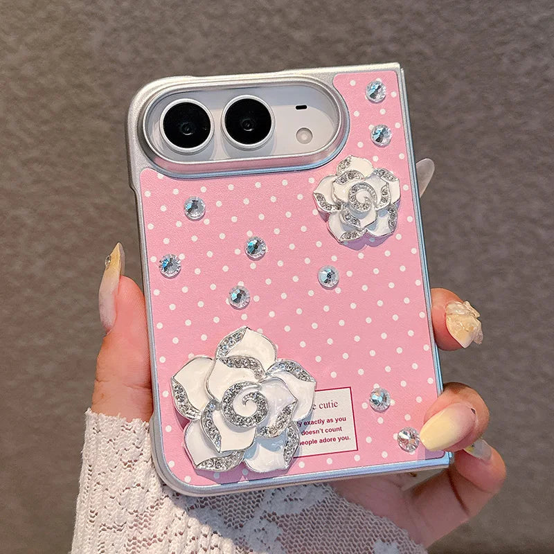 A hand holds a pink phone case decorated with white polka dots, silver rhinestones, and 3D white rose embellishments. The case has a silver bumper edge and is shown against a neutral gray textured background.