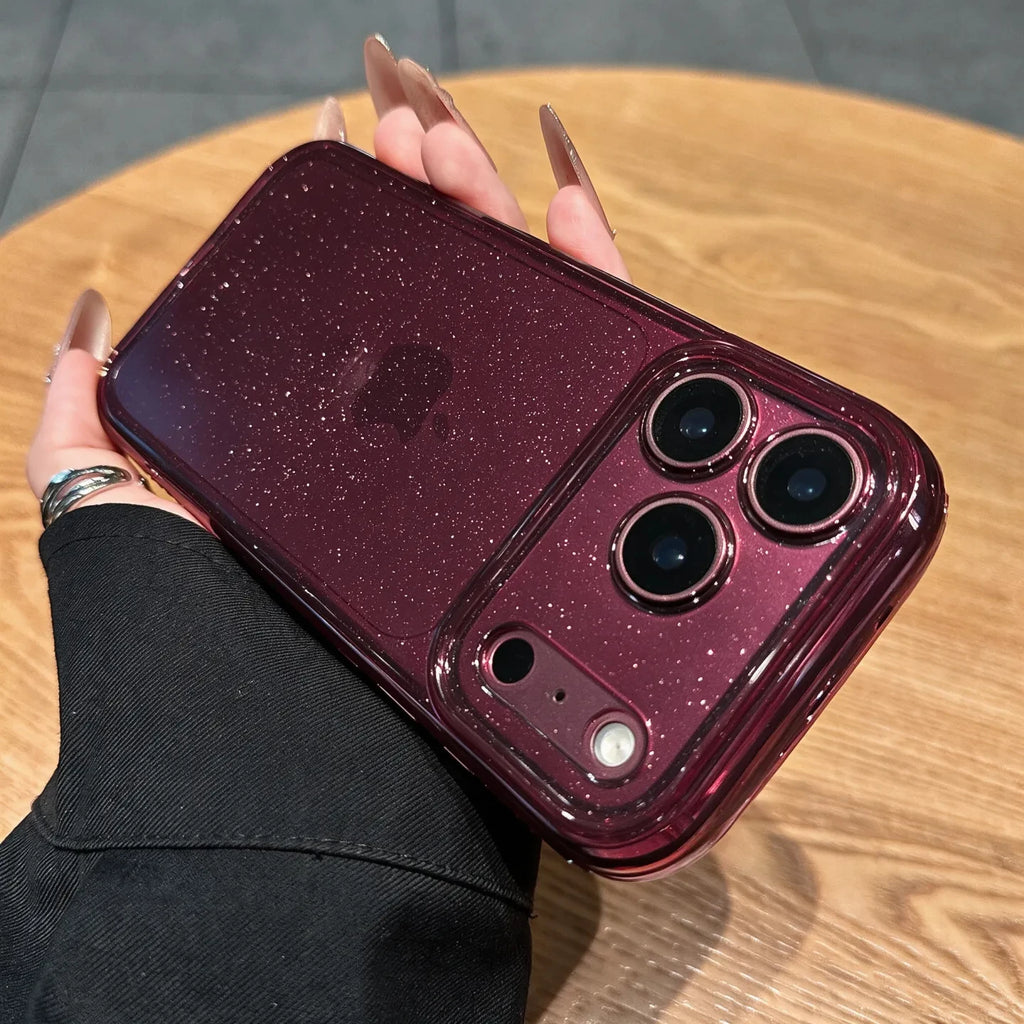 A hand with long nude manicured nails holds a smartphone in a glossy, sparkly burgundy case on a round wooden table.
