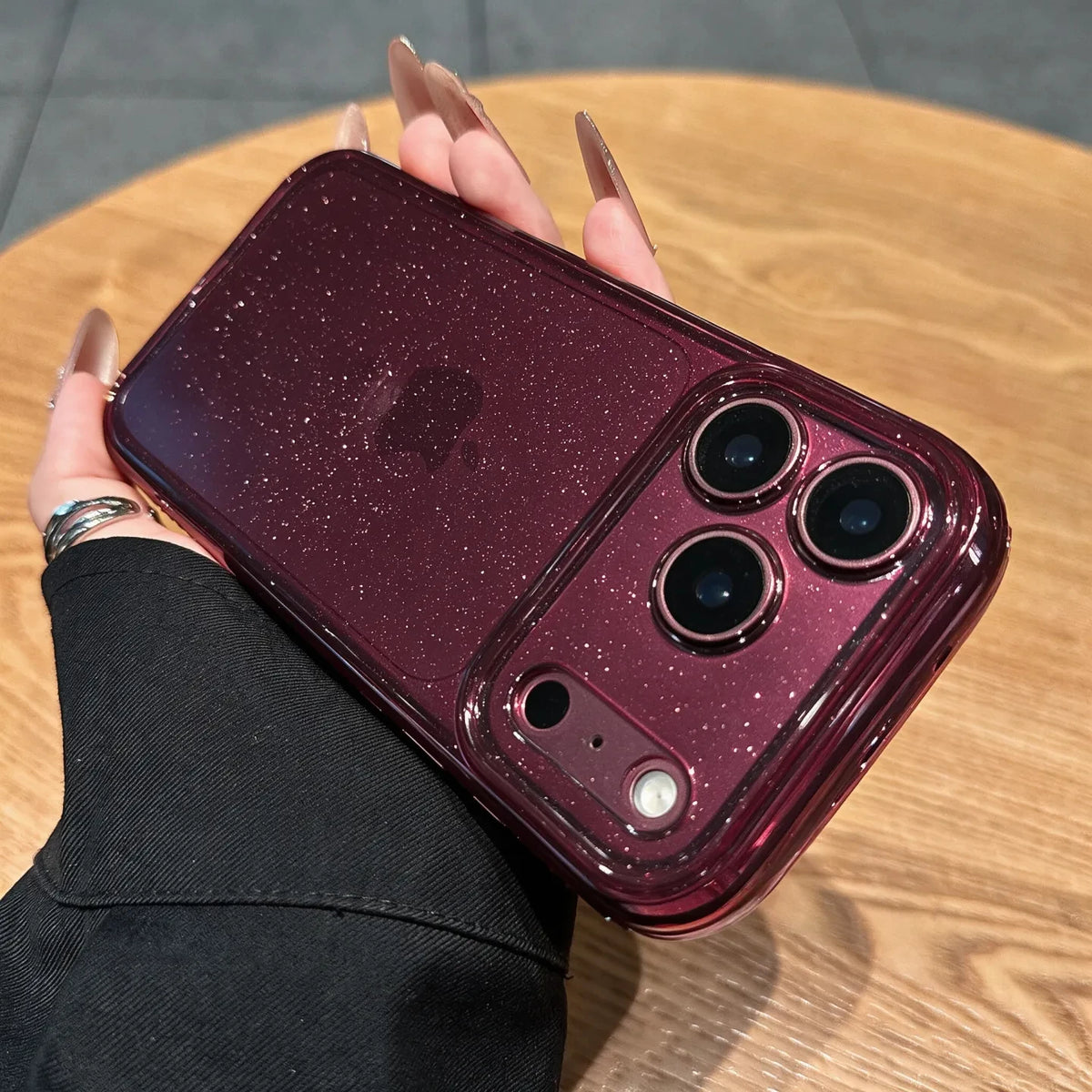 A hand with long nude manicured nails holds a smartphone in a glossy, sparkly burgundy case on a round wooden table.