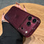 A hand with long nude manicured nails holds a smartphone in a glossy, sparkly burgundy case on a round wooden table.