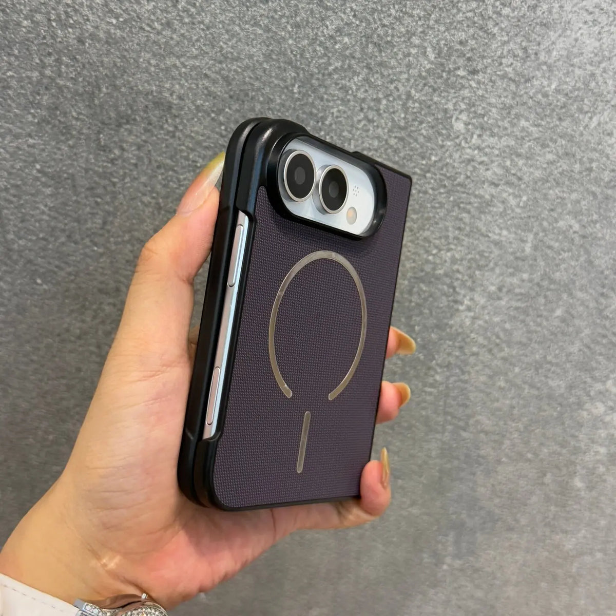 A hand holding a dark purple textured phone case with a gold MagSafe ring and a black bumper edge, set against a gray textured background.