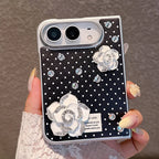 A hand with manicured nails holds a silver-framed smartphone case decorated with a black background, white polka dots, and two large white rose embellishments with rhinestone accents.