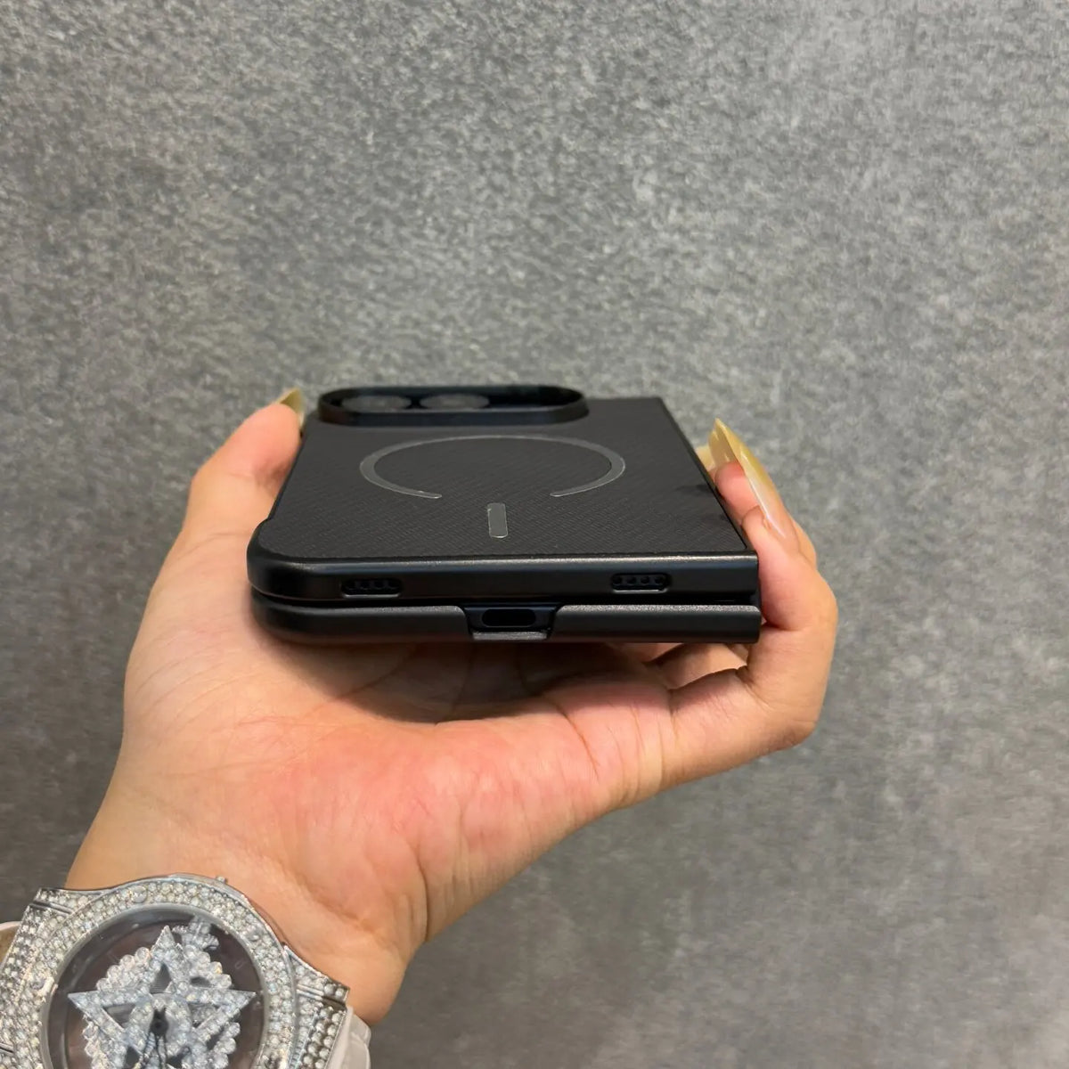 A hand holding a black magnetic phone case with a carbon fiber texture, shown against a gray textured wall. The hand has long gold manicured nails, and a sparkling silver watch is visible on the wrist.