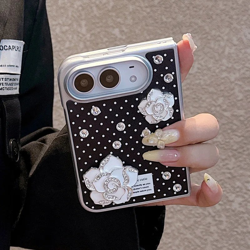 A close-up of a hand holding a black phone case decorated with white polka dots, large white flower appliqués, and rhinestone embellishments. The hand has long nude manicured nails, and the person is wearing a black jacket against a textured gray wall.
