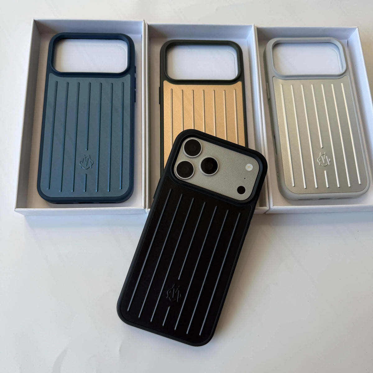 Three slim protective phone cases with vertical ridges are displayed in their white retail boxes, with one black case placed in front. The cases come in dark blue, metallic gold, and metallic silver colorways.