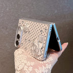 A hand wearing a cream lace sleeve holds a silver flip phone case decorated with rhinestones, polka dots, and 3D rose embellishments. The case is shown fitted on a light blue flip phone, set against a textured gray background.