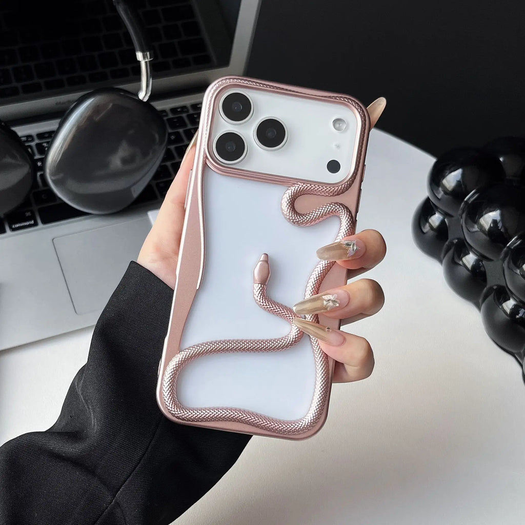 A hand holding a white iPhone case with a metallic rose gold snake design wrapping around the back, set against a desk background with a laptop and headphones.
