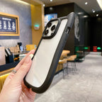 A hand holding a black and white protective phone case with a raised camera bumper, shown in a blurred cafe environment.