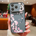 A hand holding a dark gray phone case decorated with pink and white cherry blossom flowers, shown in a blurred indoor cafe environment.