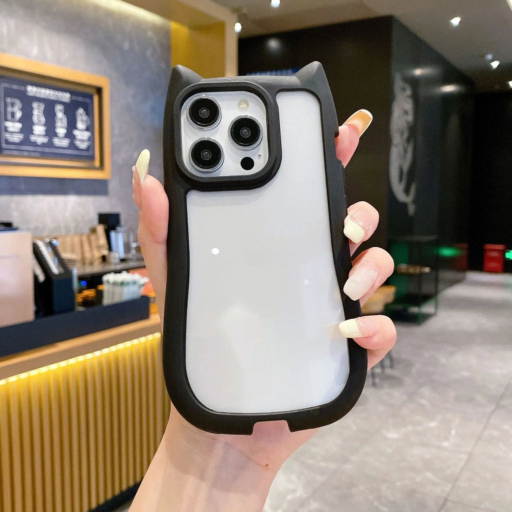 A hand holding a white phone case with a black bumper and cat-ear details, shown in a cafe setting.