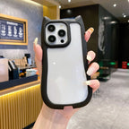 A hand holding a white phone case with a black bumper and cat-ear details, shown in a cafe setting.