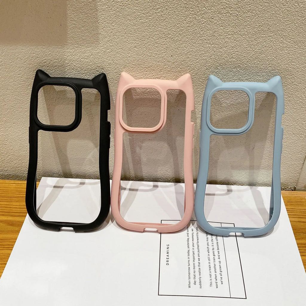 Three cat-ear phone cases in black, pink, and light blue, displayed side-by-side on a white sheet of paper against a textured beige wall.