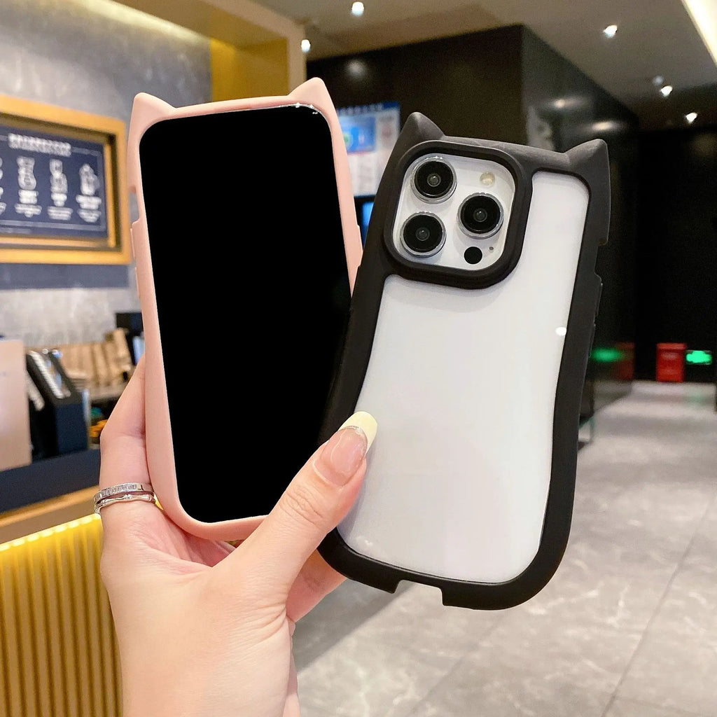 A hand holds two cat-ear phone cases in a cafe setting. One case is light pink with a black front bumper and a black phone inserted, and the other case is black with a white back and a phone inserted.