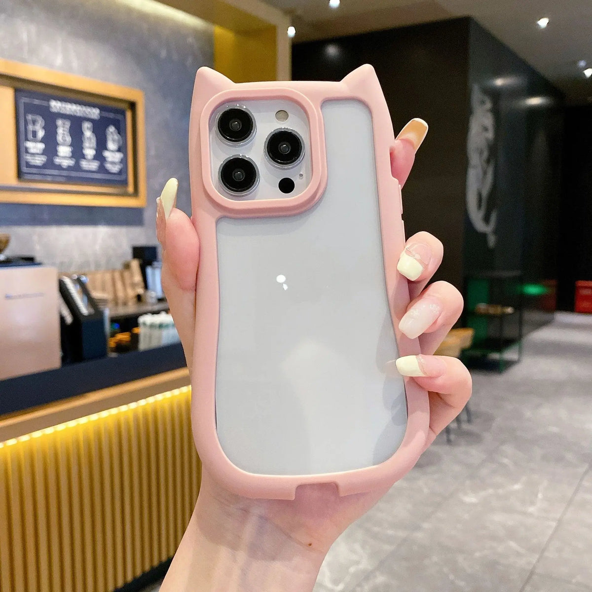 A hand holding a light pink cat-ear phone case with a frosted back panel, shown in a cafe setting.