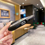 A close-up of a black protective phone case being held by a hand, showing the bottom port cutouts, with a blurred cafe interior in the background.