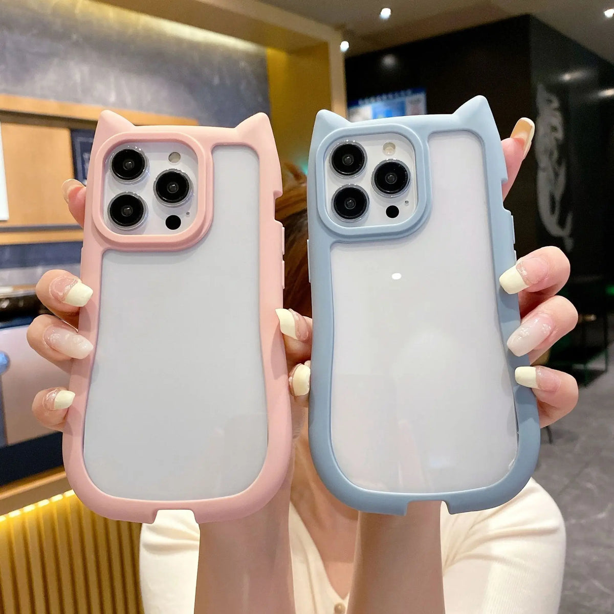 Two cat-ear phone cases held up by a person, one in pastel pink and one in pastel blue, shown against an indoor background.