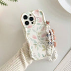 A hand holding a phone case decorated with a delicate pastel floral and leaf pattern on a white background, featuring a decorative pearl and crystal wrist strap.