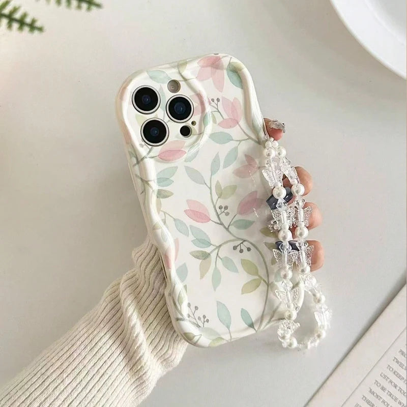 A hand holding a phone case decorated with a delicate pastel floral and leaf pattern on a white background, featuring a decorative pearl and crystal wrist strap.