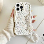 A hand wearing a cream ribbed knit sweater holds a phone case decorated with a delicate gray and green floral pattern on a white background. The case features a clear beaded wrist strap with a white bow.