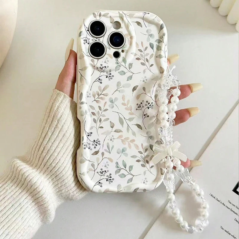 A hand wearing a cream ribbed knit sweater holds a phone case decorated with a delicate gray and green floral pattern on a white background. The case features a clear beaded wrist strap with a white bow.