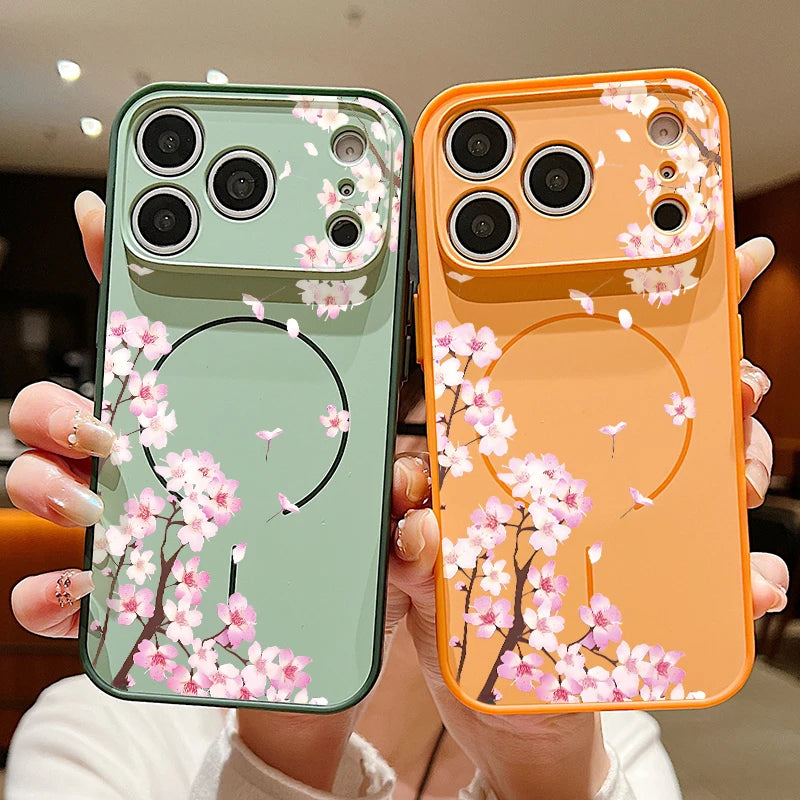 Two phone cases held in hands, one sage green and one orange, both decorated with pink cherry blossom branches and a black circular graphic.