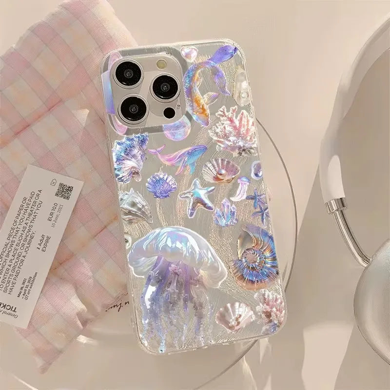 A clear phone case decorated with 3D iridescent jellyfish, starfish, and seashell designs, shown on a white phone. The case is styled on a light pink and white checkered cloth next to a product tag and a white mug.