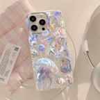 A clear phone case decorated with 3D iridescent jellyfish, starfish, and seashell designs, shown on a white phone. The case is styled on a light pink and white checkered cloth next to a product tag and a white mug.