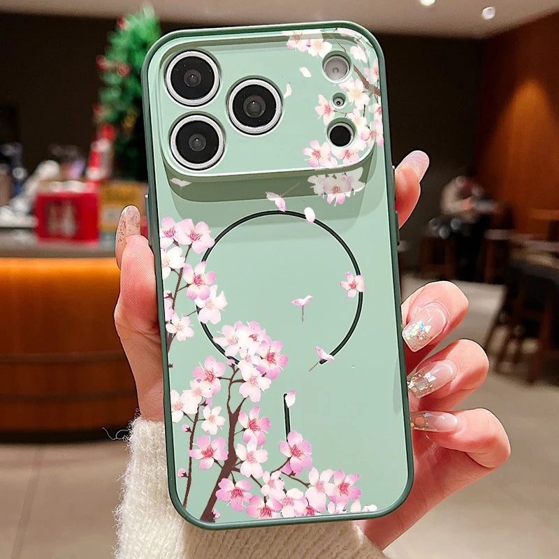 A hand holding a sage green phone case decorated with pink cherry blossom flowers and a black circular line design, set against a blurred cafe background.