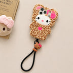 A Hello Kitty themed plush phone case with a leopard print pattern, featuring a white Hello Kitty face, pink bow, and a black wrist strap, displayed on a beige background next to a book.