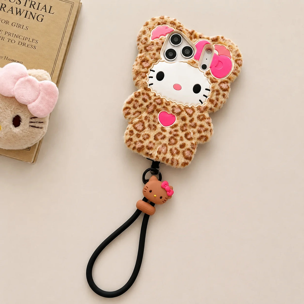 A Hello Kitty themed plush phone case with a leopard print pattern, featuring a white Hello Kitty face, pink bow, and a black wrist strap, displayed on a beige background next to a book.