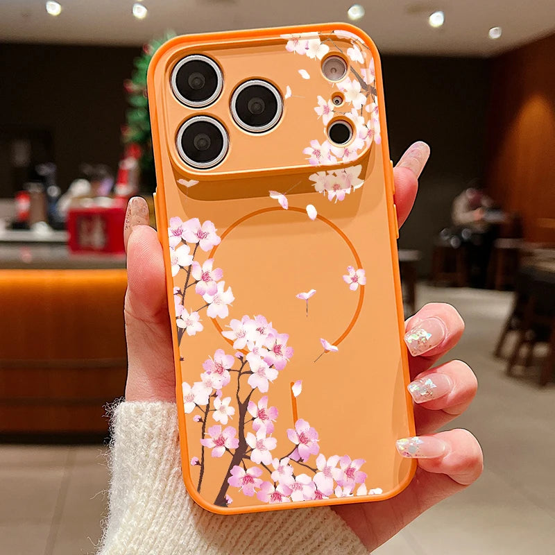 An orange phone case with a pink cherry blossom branch pattern, held by a hand in a softly blurred indoor cafe setting.
