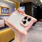 A hand holds a light pink cat-ear phone case with a clear back panel, shown against a blurred cafe background.