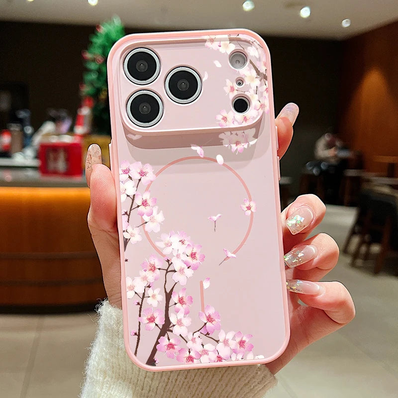 A hand holding a pink phone case decorated with cherry blossom branches and a large circular frame design, photographed in a blurred indoor cafe setting.