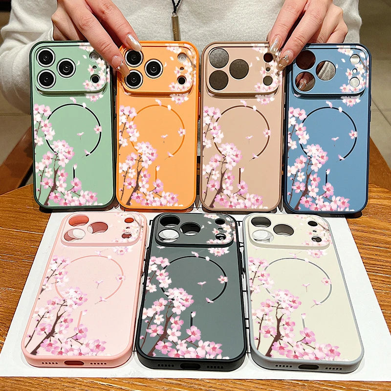 Seven phone cases with a pink cherry blossom branch pattern are displayed, held by hands over a wooden table. The cases come in various colors including sage green, orange, champagne beige, sky blue, light pink, dark gray, and off-white, each featuring a circular magnetic ring design.