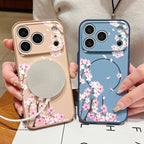 Two floral smartphone cases held in hands, one in a soft blush pink and one in a dusty blue, both printed with pink cherry blossom branches and a circular MagSafe-style magnetic ring.