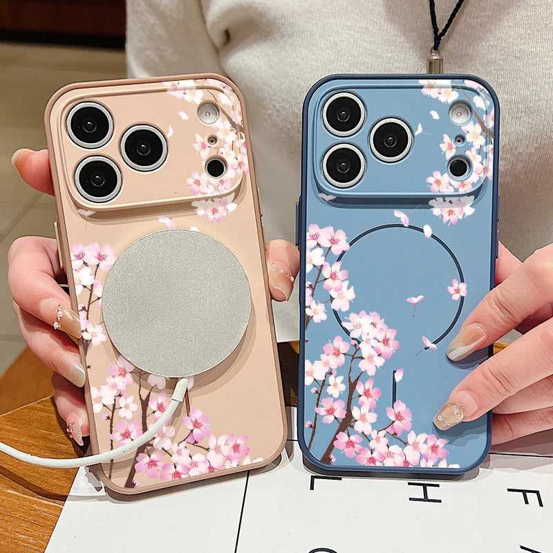 Two floral smartphone cases held in hands, one in a soft blush pink and one in a dusty blue, both printed with pink cherry blossom branches and a circular MagSafe-style magnetic ring.