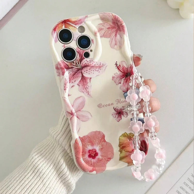 A hand wearing a white ribbed knit sleeve holds a phone case decorated with pink and red hibiscus flowers on a cream background. The case has a clear beaded wrist strap with pink flower charms.