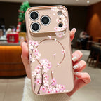 A hand holding a phone case decorated with pink cherry blossom flowers and brown branches on a light beige background, set against a blurred cafe interior.