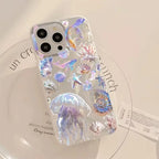 A clear phone case decorated with iridescent jellyfish, seashells, and starfish, shown on a white phone resting on a white plate.