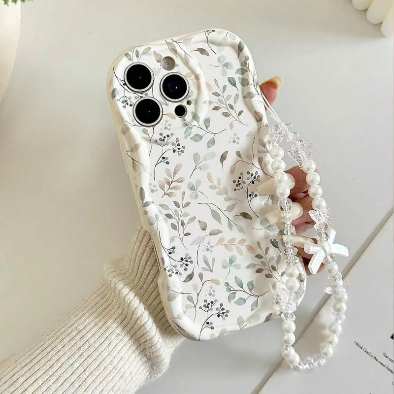 A phone case with a delicate white, gray, and beige floral pattern, held by a hand wearing a cream-colored ribbed sweater. The case is adorned with a decorative pearl bead wrist strap featuring small white bows and clear rhinestone accents.