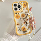 A hand holding a phone with a floral yellow and white phone case featuring a pearl strap, set against a light background.