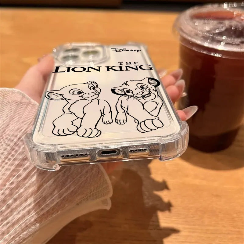 A hand holding a clear phone case featuring black line art illustrations of Simba and Nala from Disney's The Lion King, with the movie title printed above. The hand is resting on a wooden table next to an iced coffee.